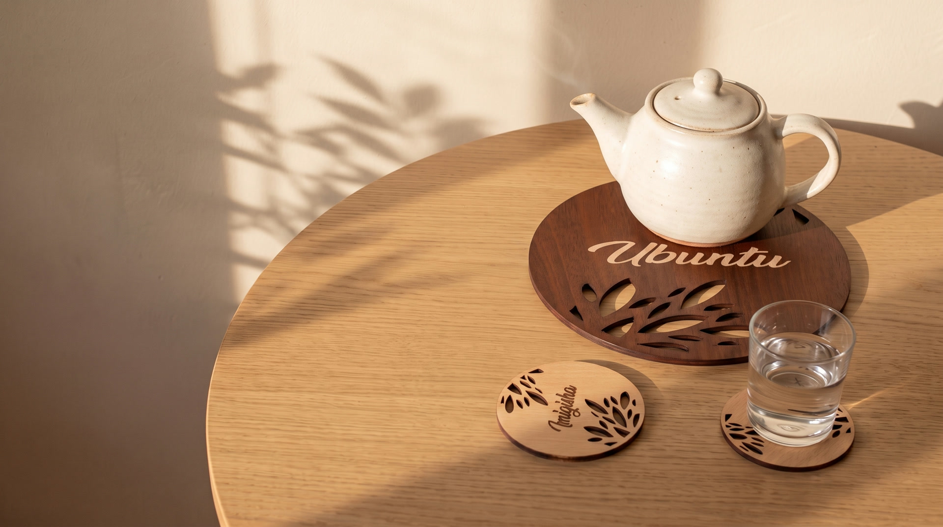 Une table en chêne au soleil : une théière posée sur le sous-plat Ubuntu, un sous-verre Imigisha à côté d'un verre d'eau